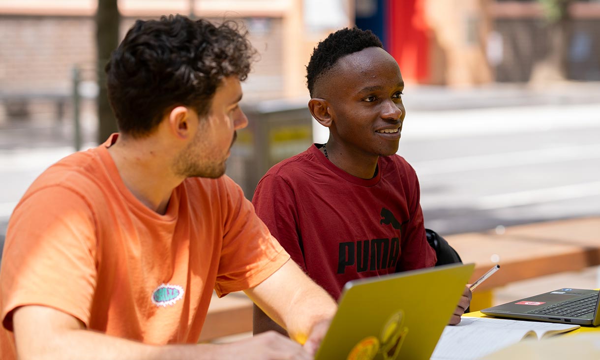Two students in orange and red tshirts sitting with laptops outseide.