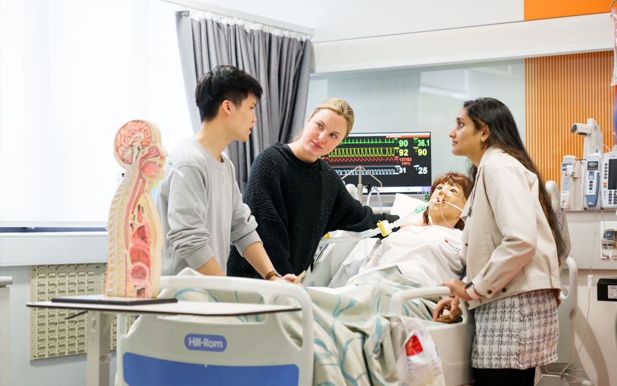 Three students standing in a medical simulation room with a patient manikin