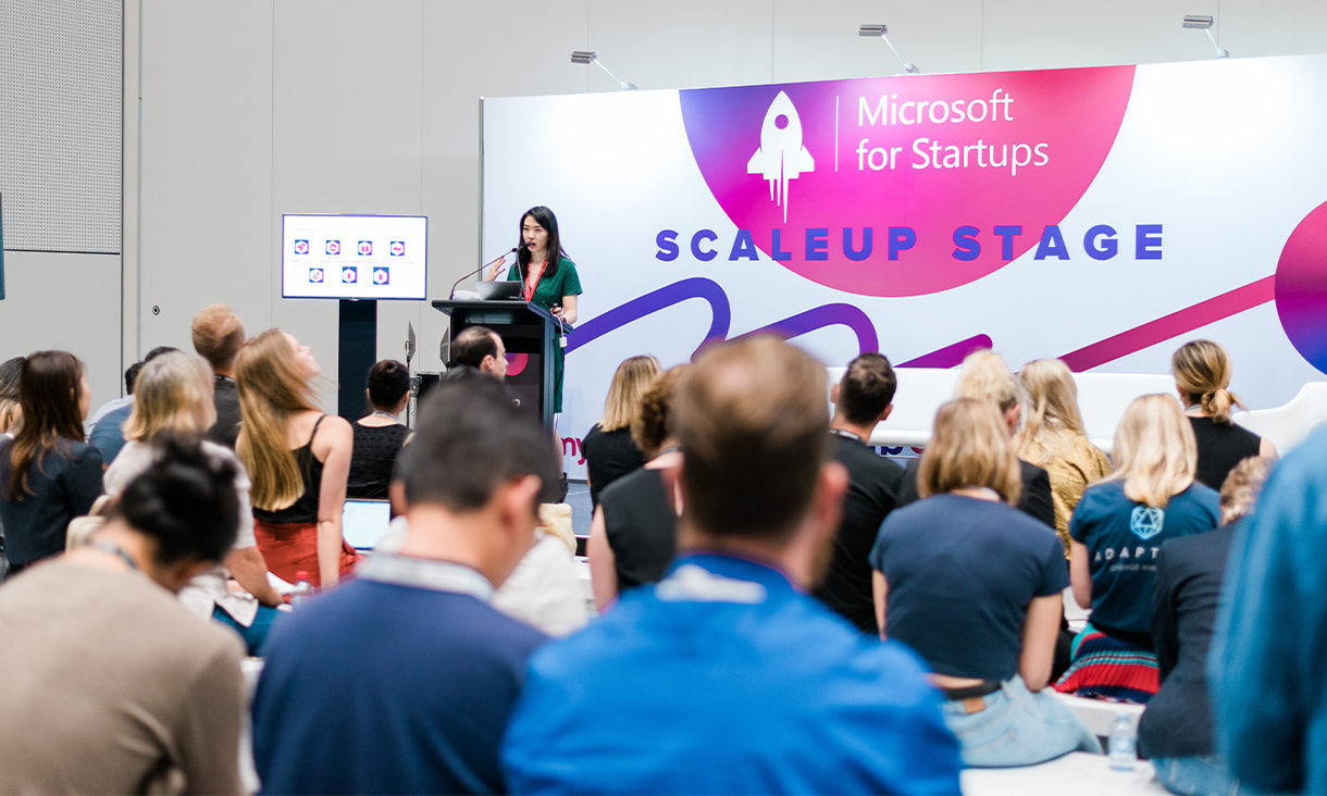 RMIT alumni standing in front of a podium, addressing an audience, in front of a wall, labelled "Microsoft for Startups: SCALEUP STAGE"