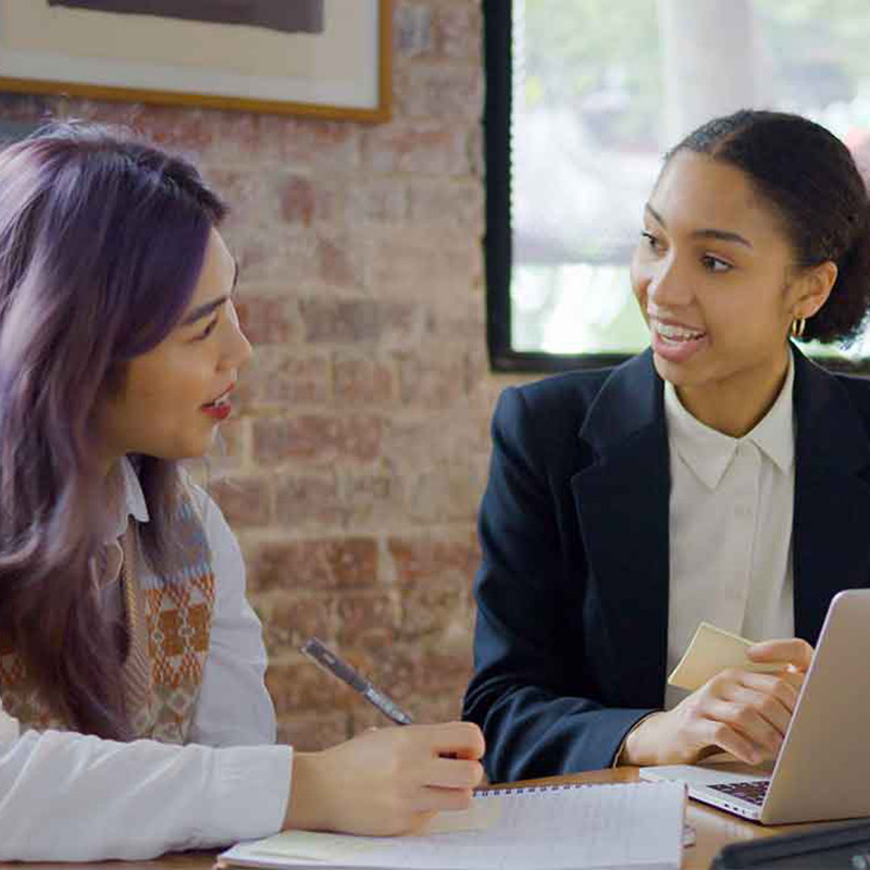 Two female students studying together at a desk, one with a book and pen, the other with a laptop.