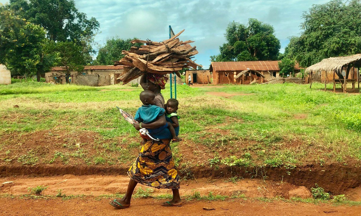 Woman in a bright skirt carrying wood and two small children outside
