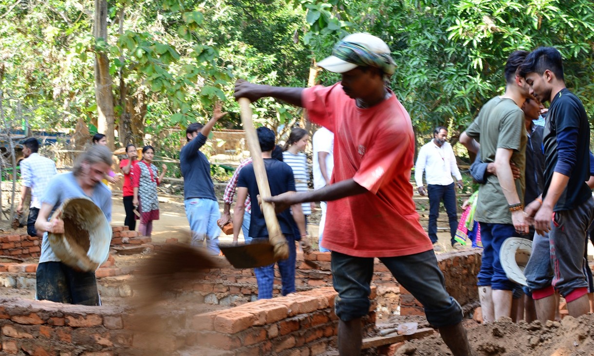 Several students laying bricks and building a mud house outdoors