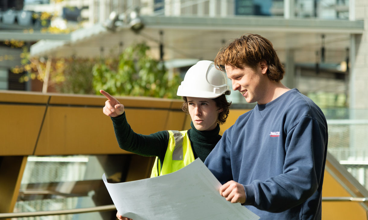 Female student wearing hard hat, pointing, while male student is holding large draft plan in front of them