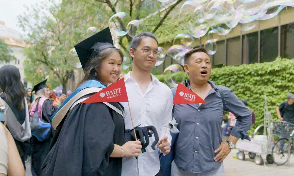 RMIT 2023 graduate wearing a graduation gown and cap, holding a red RMIT flag, standing with her siblings on Bowen Street, after her graduation ceremony