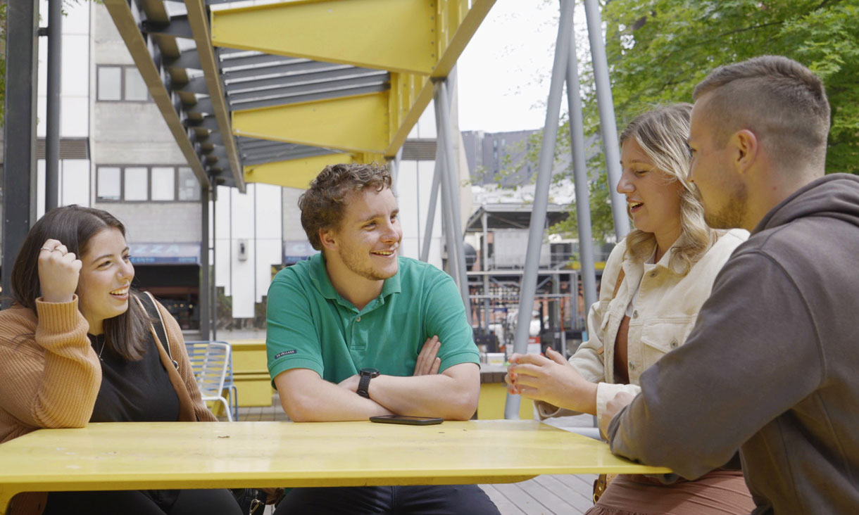 Thomas, sitting at a table, surrounded by student friends, on the RMIT city campus, outside on Bowen Street