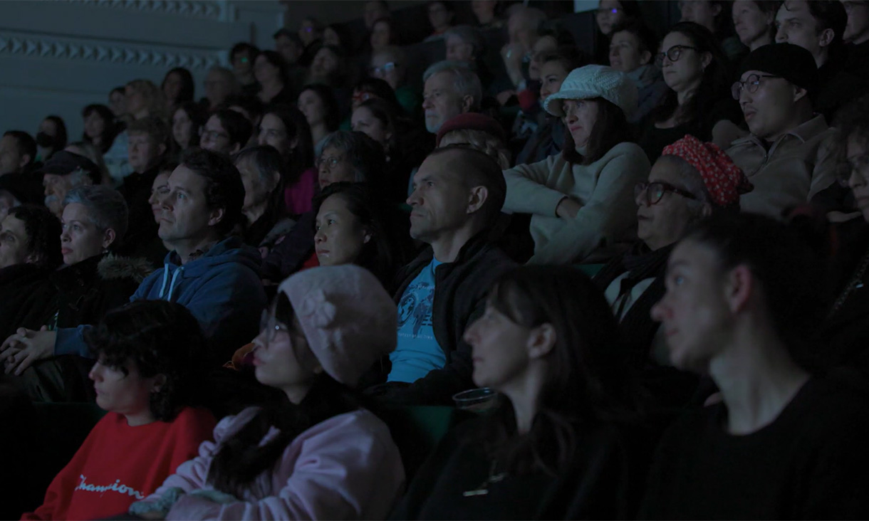 A group of people is seated in a dimly lit auditorium. The audience appears to be watching attentively, with some individuals wearing casual winter clothing, such as jackets and sweaters. 