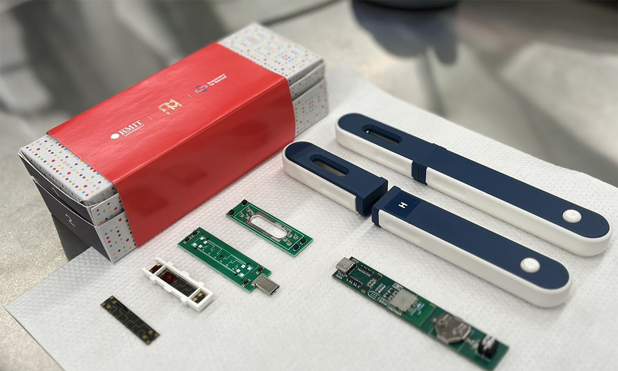 The integrated Wireless Biosensor Device laid flat on a stainless-steel bench. It is broken down into its components so we can see how it is made. It is navy blue and the size of a travel toothbrush case. There is an example of the package design at the back.