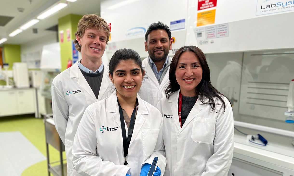 Two male researchers are standing behind two female researchers in the lab. They are all wearing white lab coats and one of the women is holding a testing device.