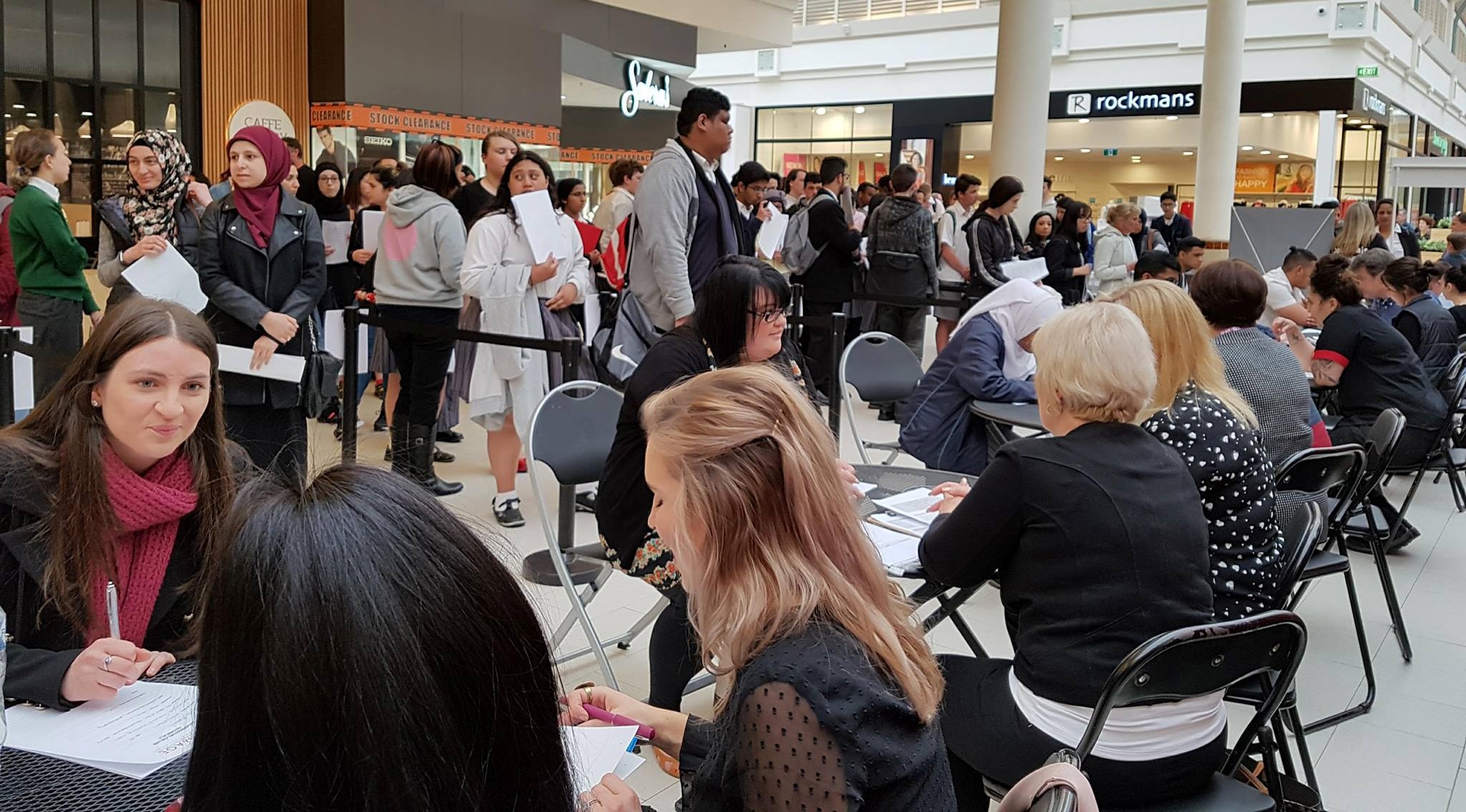Photo taken inside a shopping centre. Foreground shows people sitting at separate tables, some writing on pieces of paper. In the background, people are forming a queue.