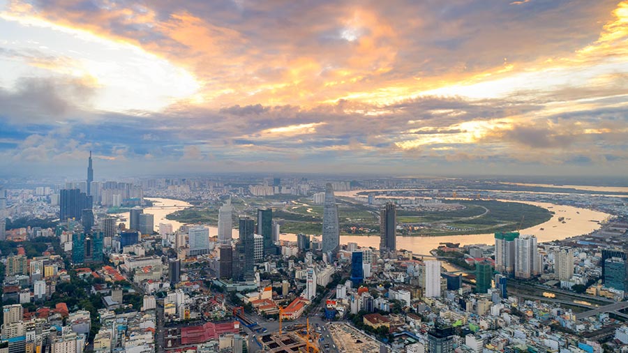 Aerial photo of Ho Chi Minh City. Sun is setting with clouds shining a bright yellow to orange gradient. The river surrounding the city reflects the orange colour from the sky.