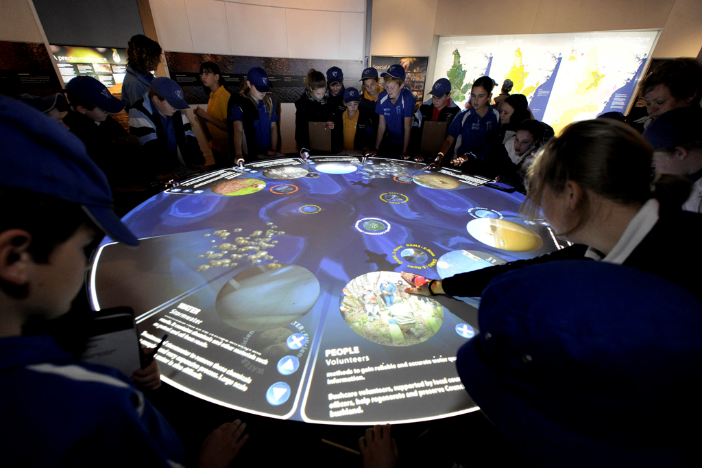 Photo of a circular digital display on a table. People are surrounding the table and one person is pointing at one point on the digital display.