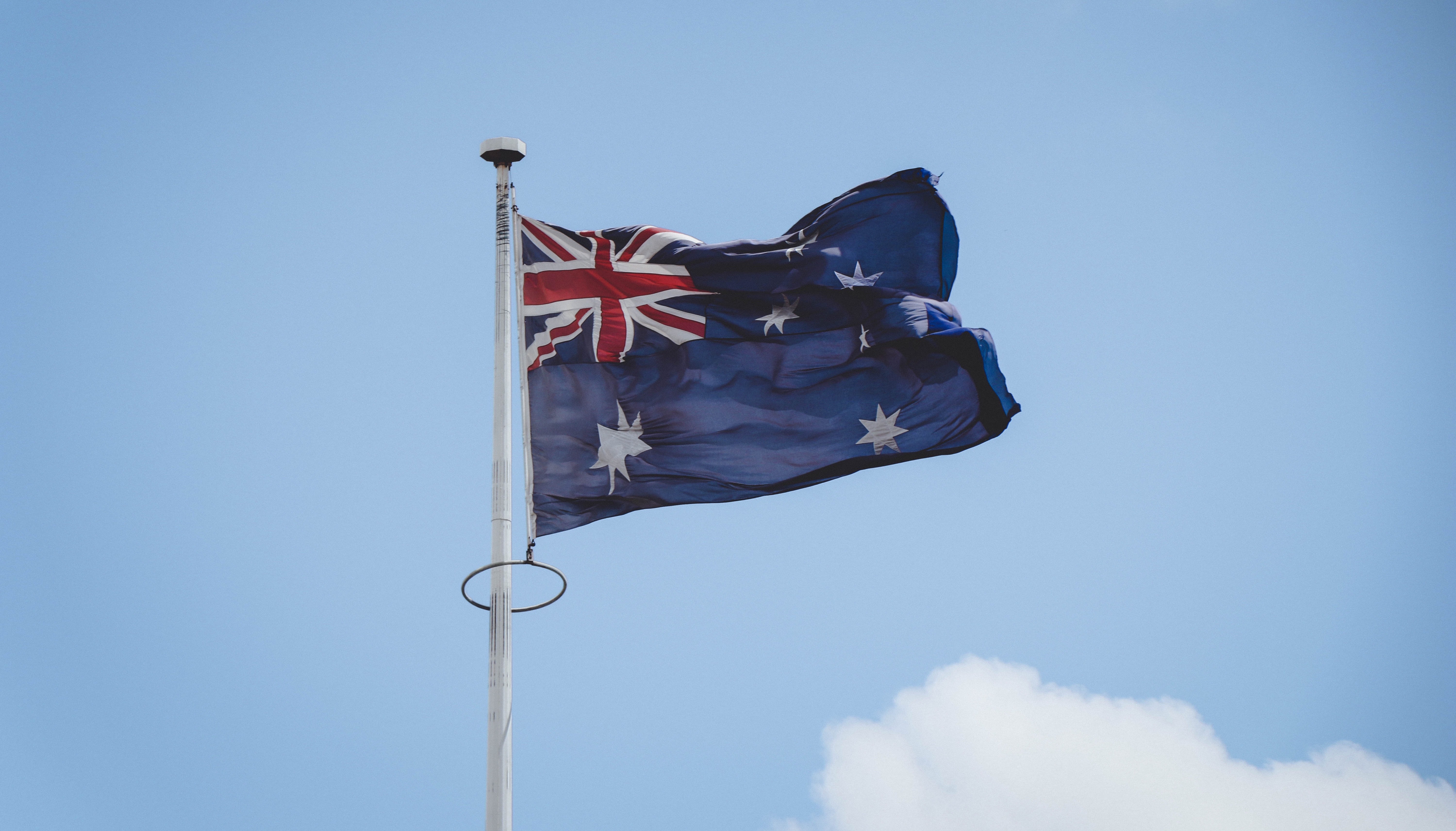Photo of the Australian flag blowing in a strong breeze, attached to a metal flagpole. Background is a blue sky with clouds forming at the bottom of the photo.