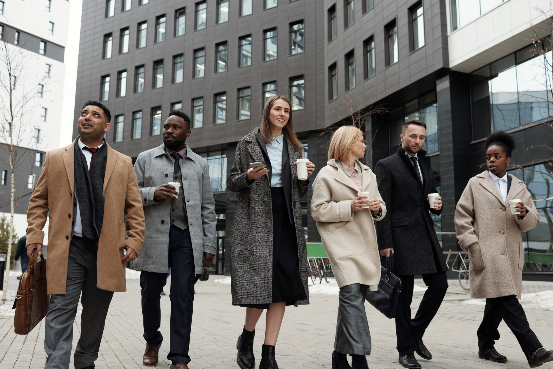 Group of six people working in a city. Five of the people are holding coffees. Everyone is wearing a coat and walking in the same direction. They're looking in different directions.