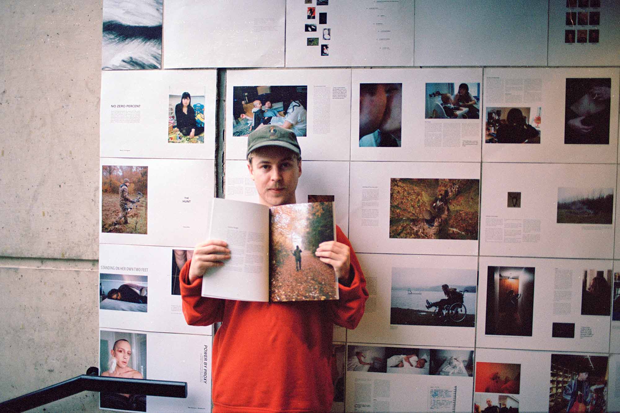 Man holding up open book, standing in front of a wall of photos 