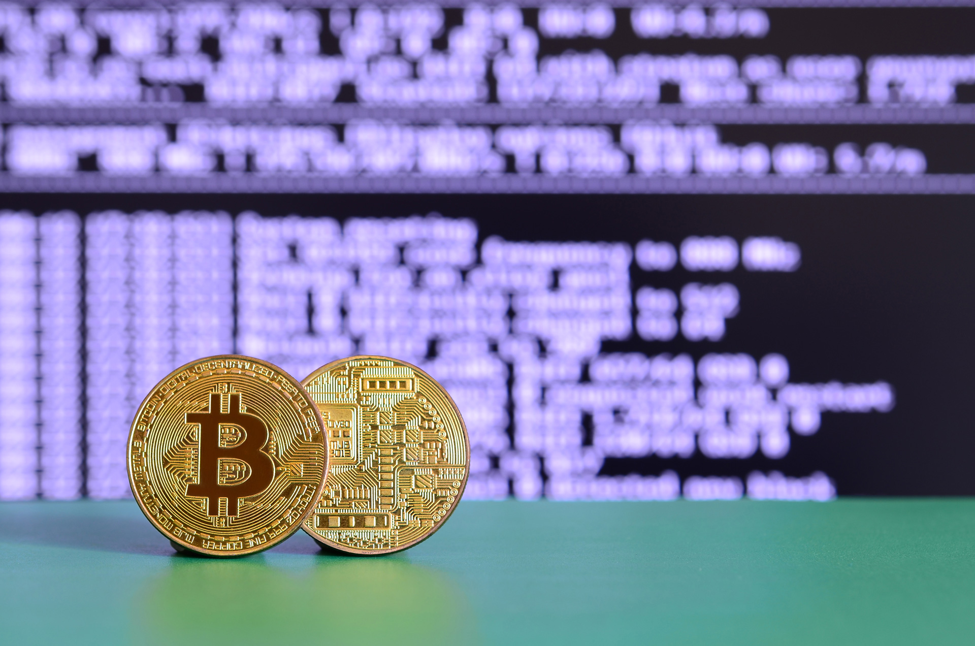 Two gold bitcoins lie on the green surface on the background of the display, which shows the process of mining the crypto currency