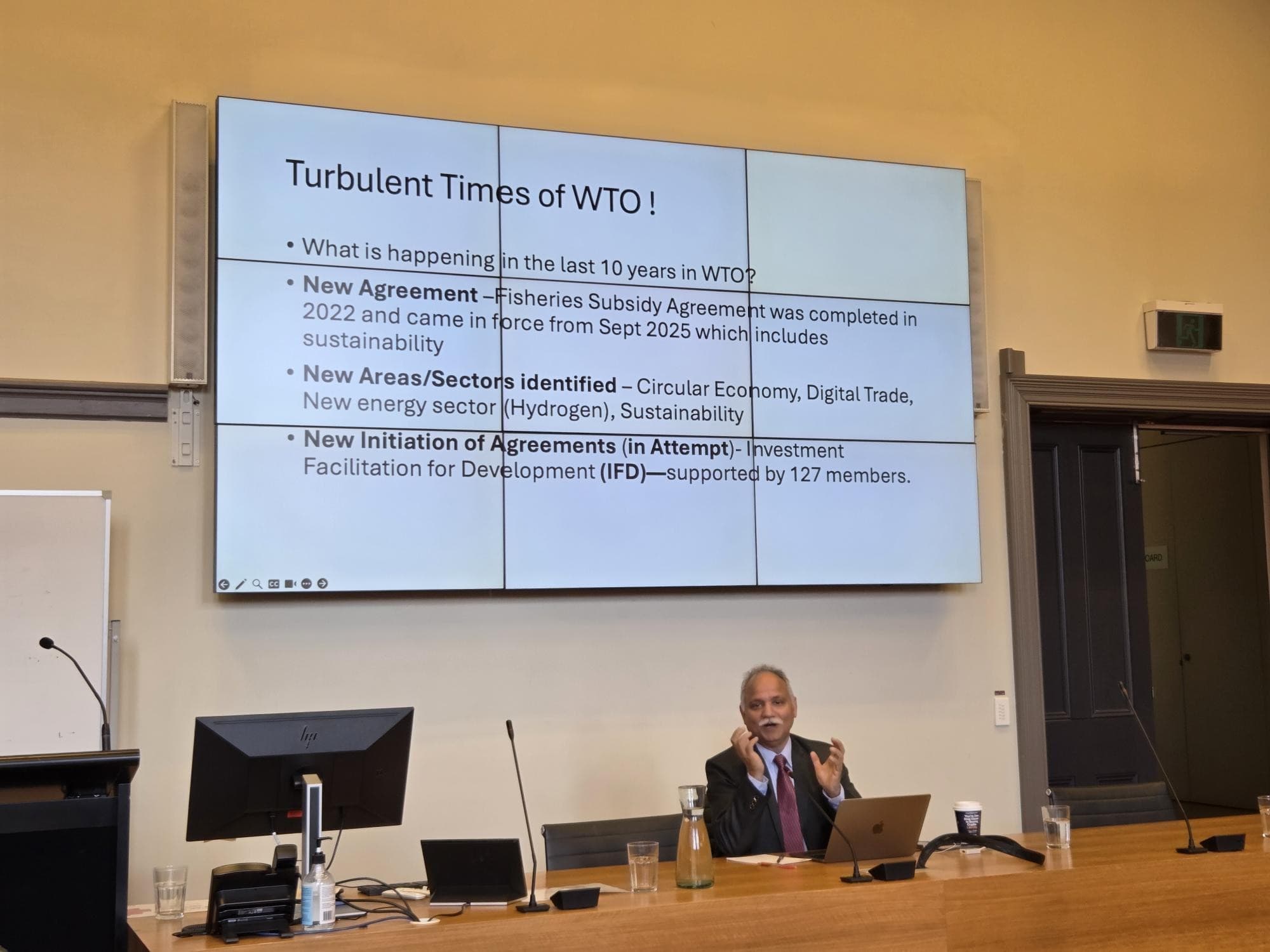 Speaker in front of presentation titled Turbulent Times of WTO!
