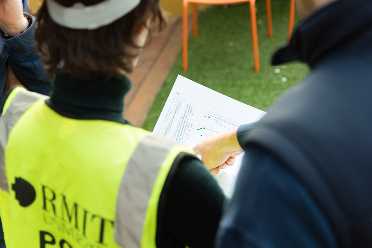 Student wearing RMIT hi-vis safety vest as instructor points towards a list, held by student.