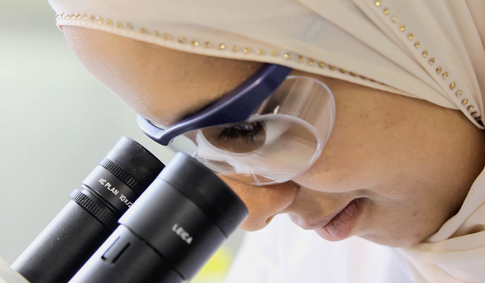 Female wearing safety goggles, looking into microscope lens