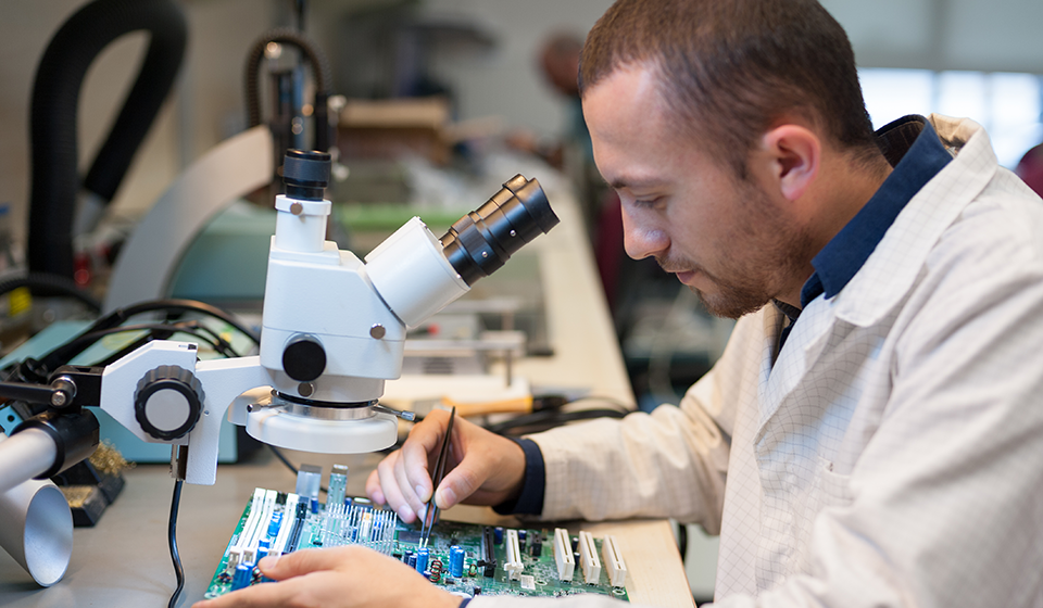 A male wearing a white lab-coat, sitting in front of a microscope, working on an electronic circuit board