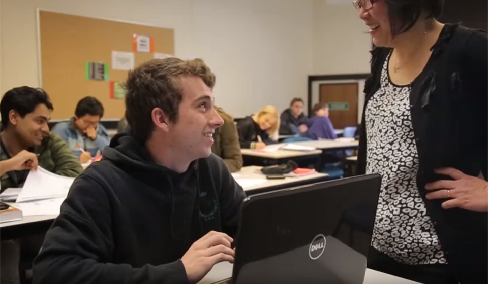 Male student in front of laptop, talking to teach standing in front of him, inside a classroom