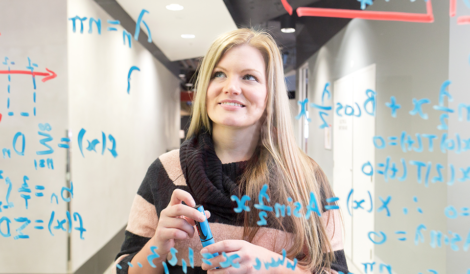 Femalre student standing in front of clear glass writing board with complex written formulas