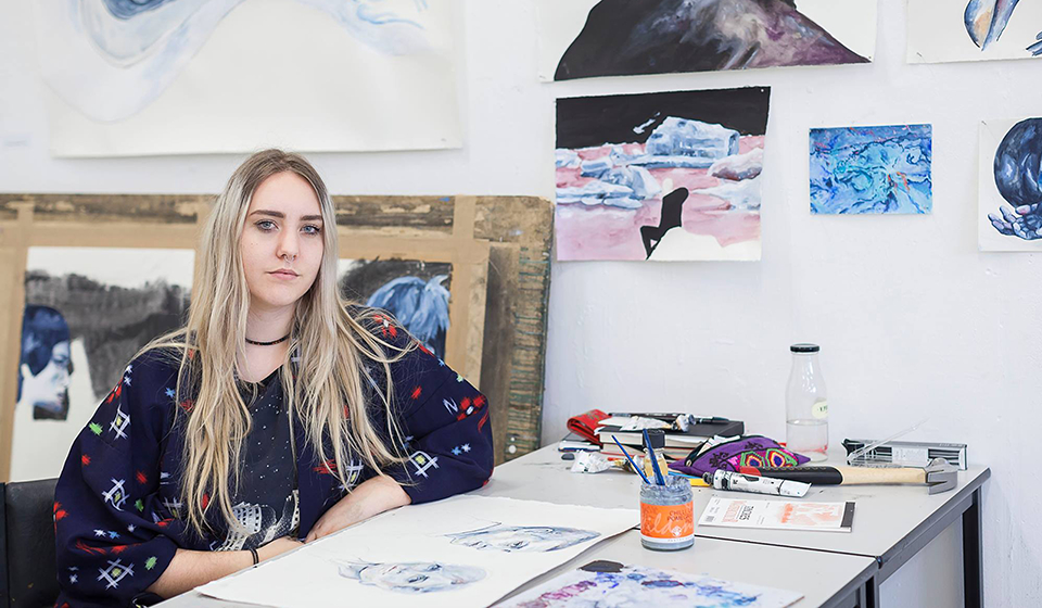 Female student resting arm on desk in studio, surrounded by artwork on walls and desk