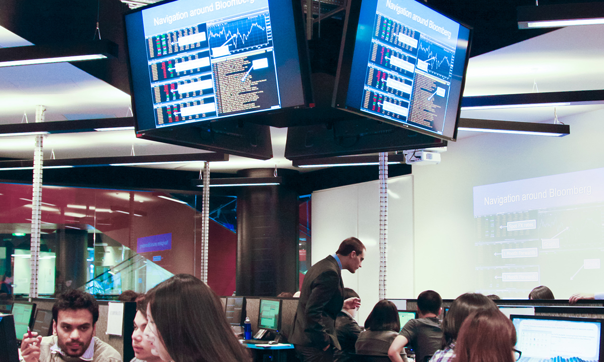 Trading facility room filled with students, with focus on screens connected from ceiling displaying graphs