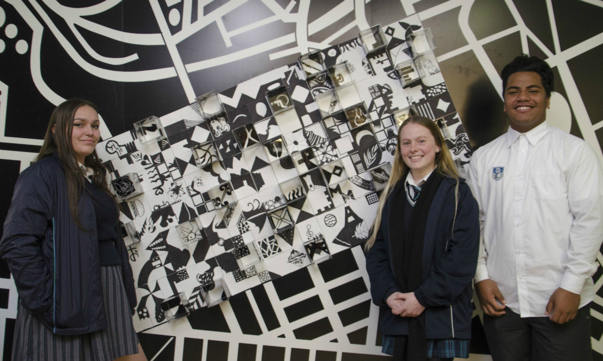 Three smiling students stand in front of their mural on the wall. 