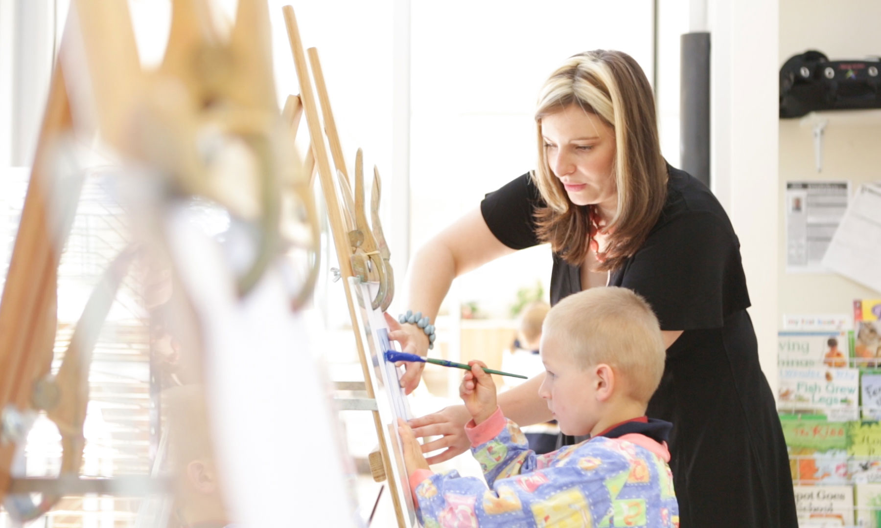 A classroom teacher assists a child painting at an easel.