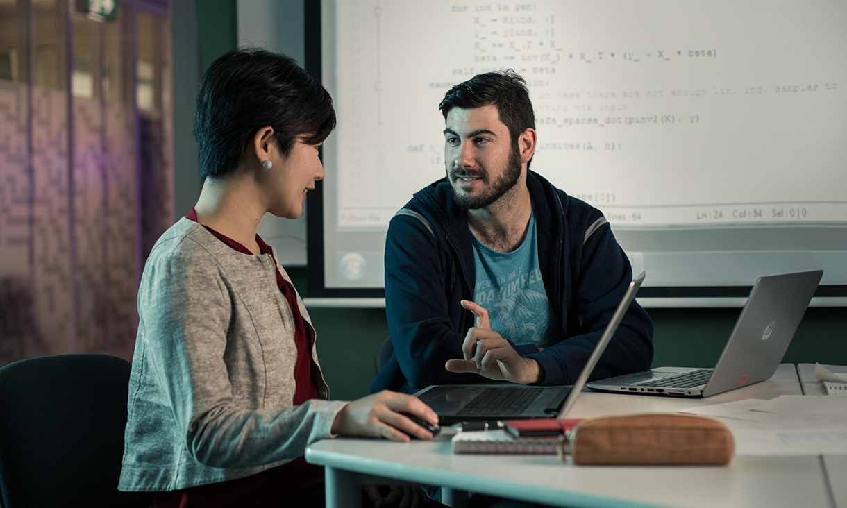 A male and female student sitting at a desk, both with open laptops, discussing an IT project