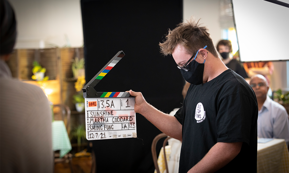A student wearing a black cloth face mask holds a clapperboard on a film set.