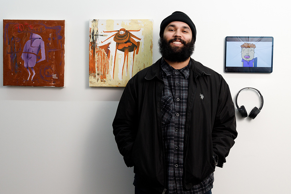 Artist Tyrown Waigana in front of 3 illustrations at the Lamington Drive gallery space