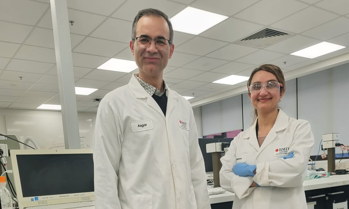 Two RMIT scientists stand in their lab, wearing white lab coats, blue disposable gloves and protective goggles. They are smiling at the camera.
