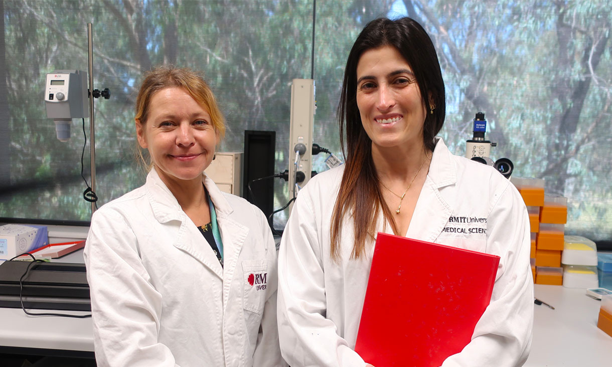 Dr Céline Valéry with PhD candidate Priscila Cardoso wearing lab coats in their science lab. Priscila is holding a red folder.