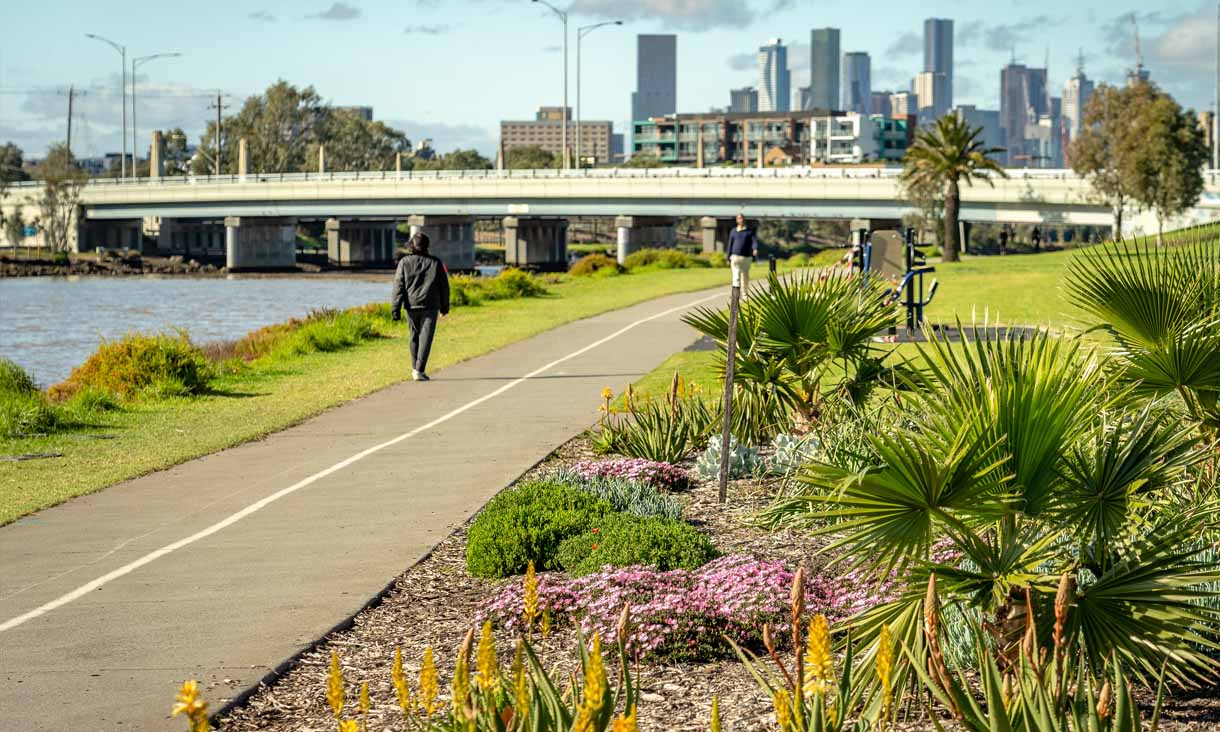 A person is walking along the Maribyrnong River. The path is surrounded with plants. A bridge is visible in the distance.