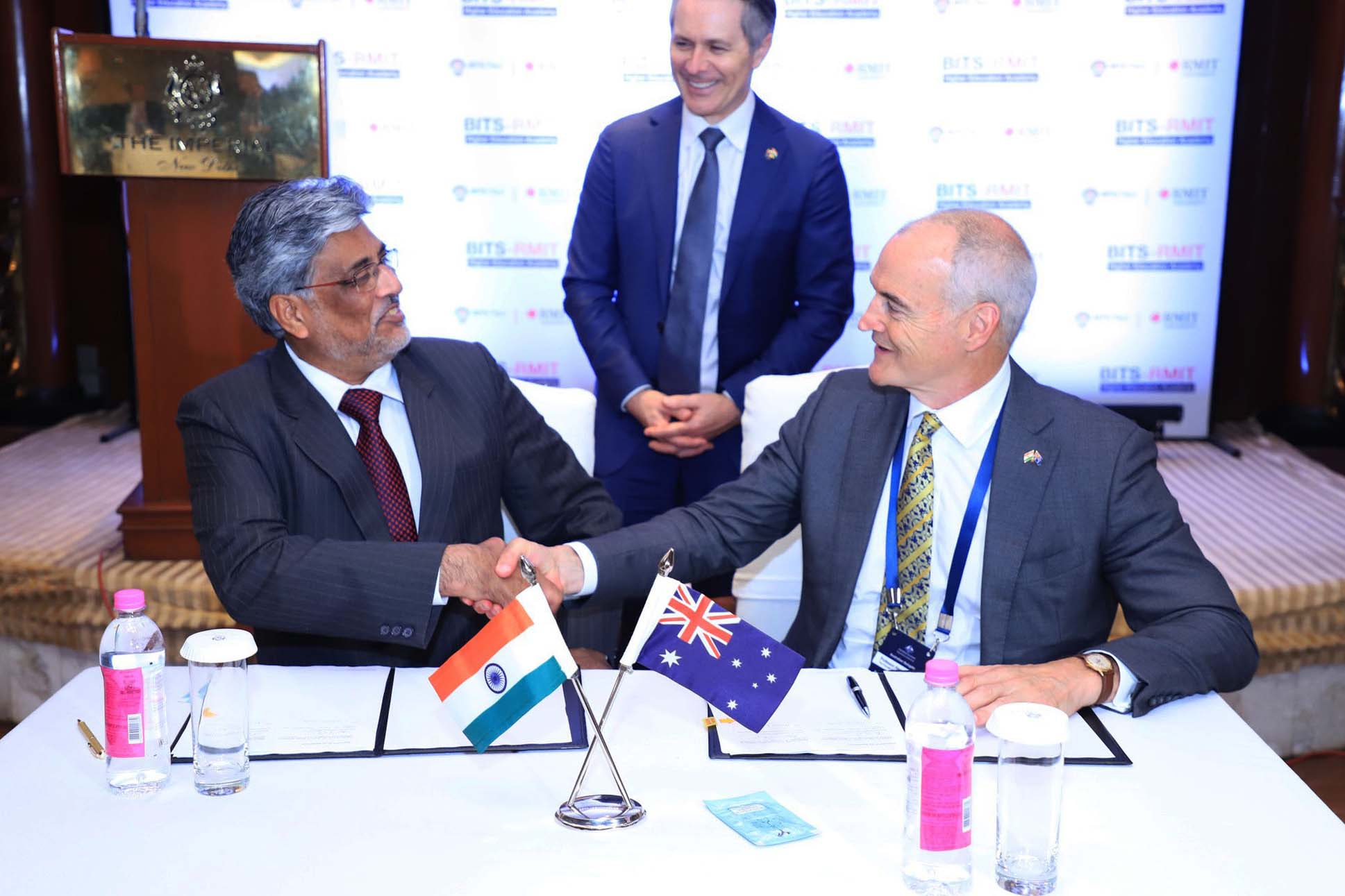 Prof Souvik Bhattacharya BITS Vice-Chancellor and Prof Alec Cameron RMIT Vice-Chancellor and President shake hands while sitting at a desk. Hon. Jason Clare MP Federal Minister for Education watches and smiles.
