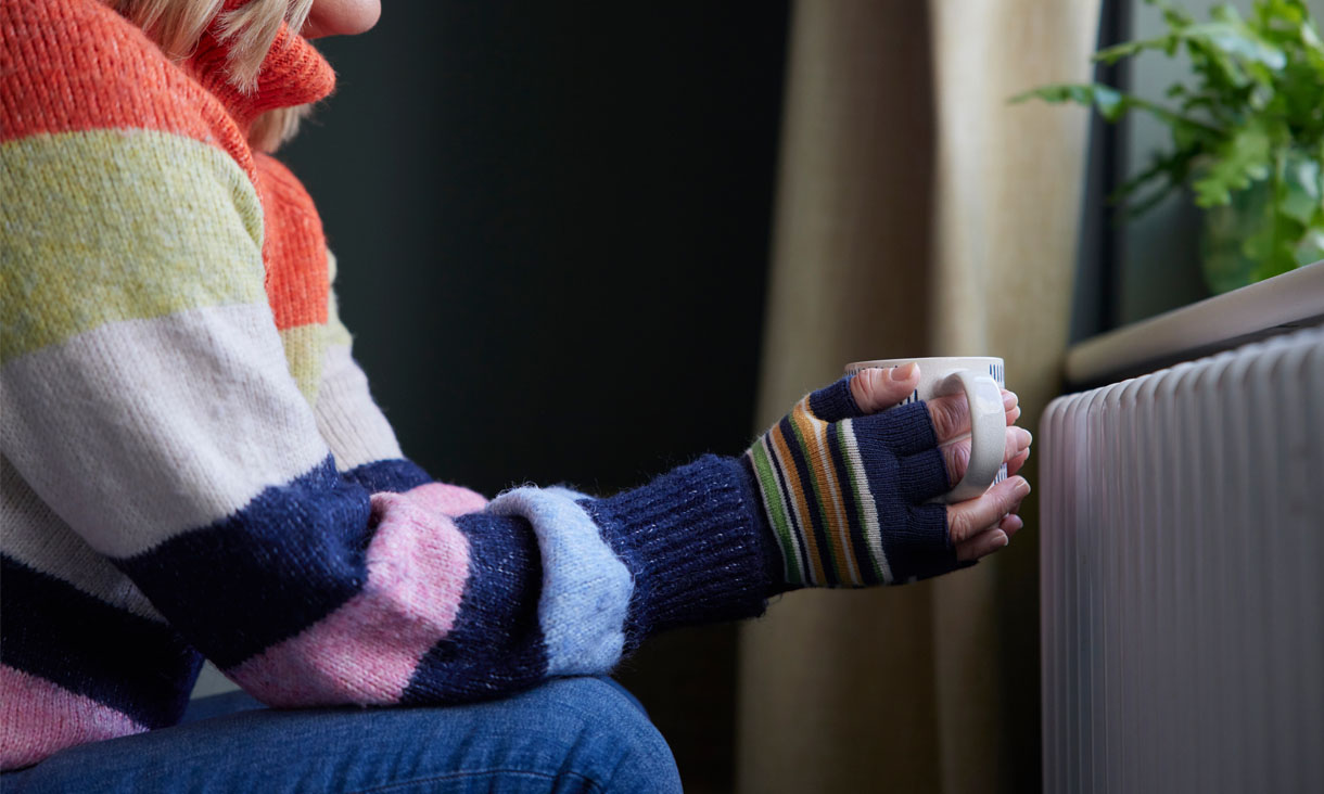 A person wearing a thick jumper and gloves tries to keep warm by holding a mug while sitting in front of a heater. 