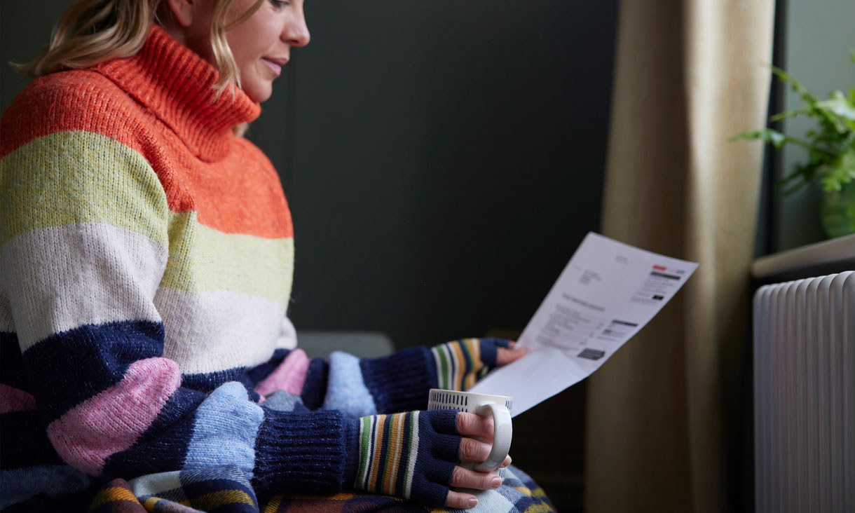 A person wearing a thick jumper and gloves sits in front of a heater, holding a mug and looking at an energy bill.