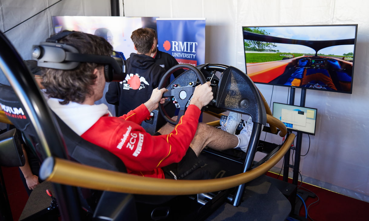 A boy sits in a model of a racecar with a VR headset on. The screen in front of him shows the inside of a car on a racetrack. 
