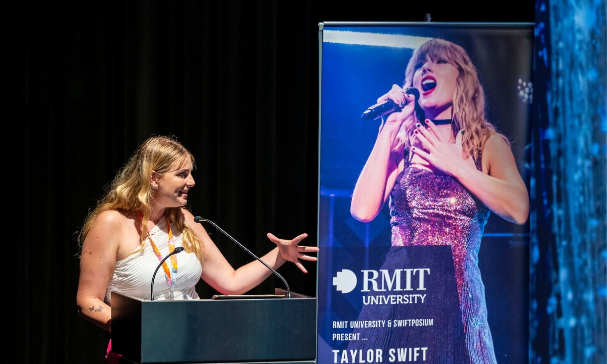 Jasmine presenting at lectern, with Taylor Swift banner behind her