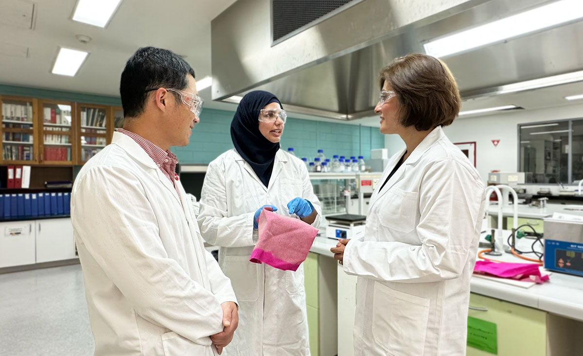 From left to right: Research supervisor and RMIT Senior lecturer Dr Xin Wang, Lead researcher and RMIT research assistant Dr Aisha Rehman and Project leader and RMIT Senior Lecturer Dr Shadi Houshyar. Aisha is talking and holding pink cotton that's been treated with nanodiamonds while Xin and Shadi listen to her. Everyone is wearing white lab coats and protective goggles. 