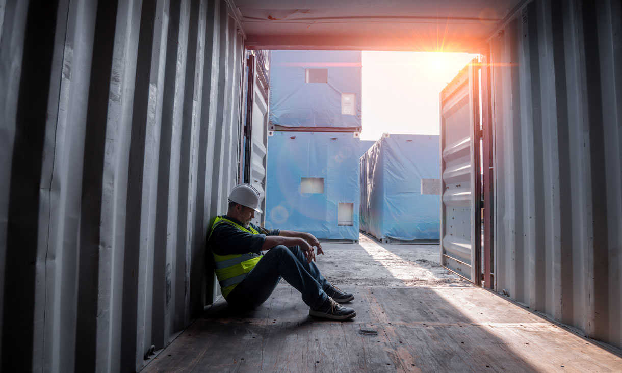 A construction worker in a hard hat and high vis vests sitting in a shipping container looking sad.