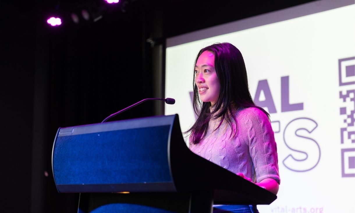 Claire Qian is photographed speaking and smiling at podium under purple lights. 