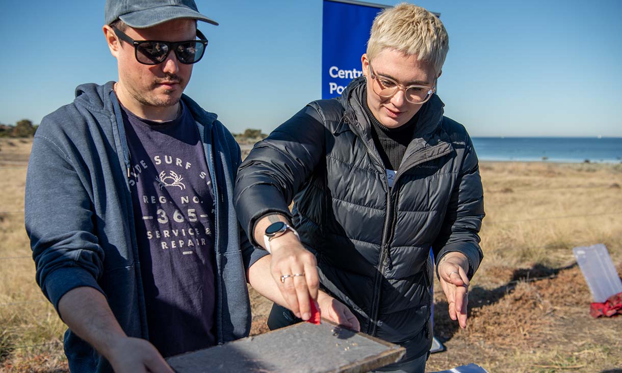 Two individuals collaborate on an outdoor task, one holding a frame while the other applies material, with a blue banner in the background.