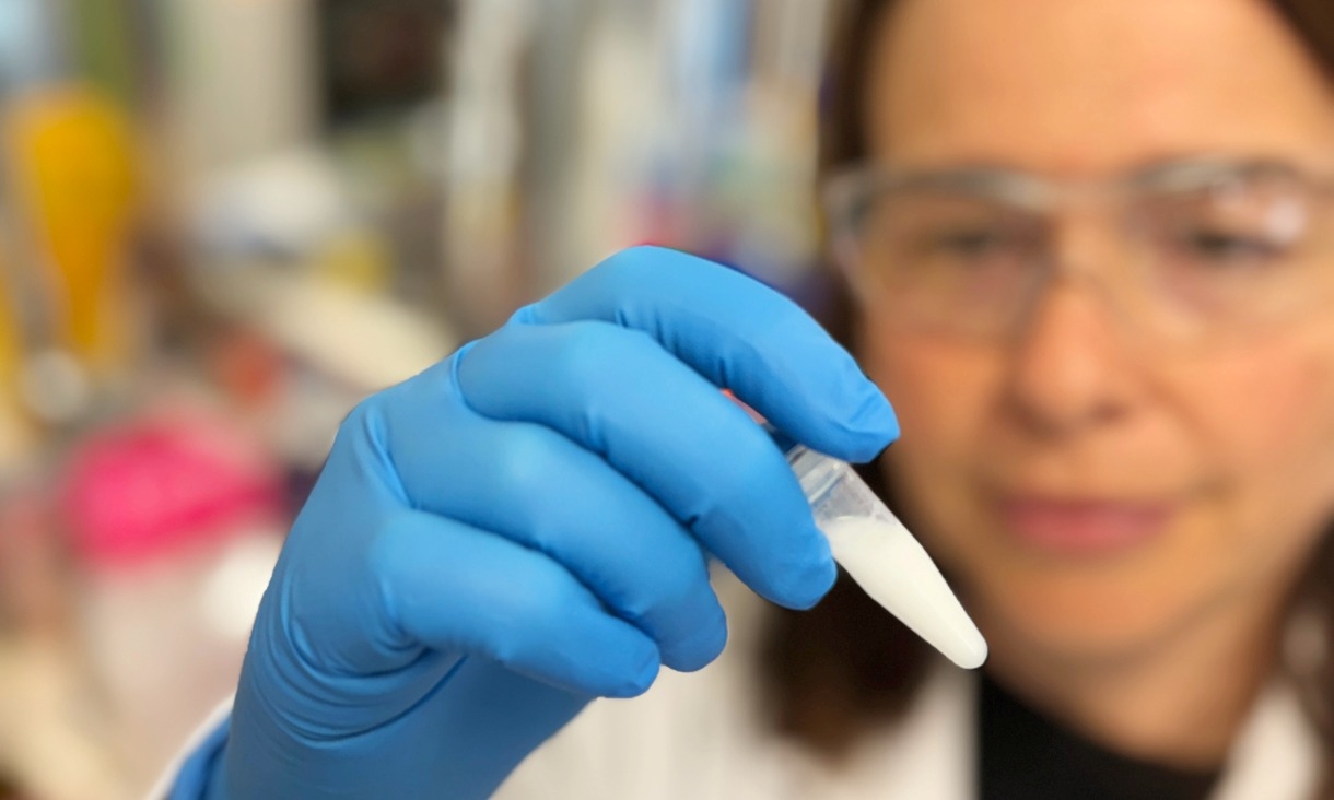 A woman in a white lab coat and safety glasses holds up a vial of white liquid in a laboratory.