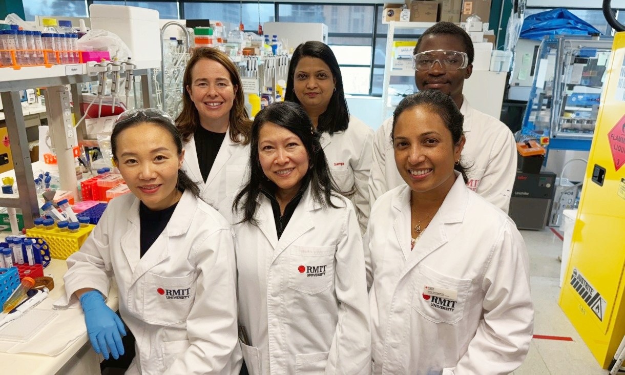 Six researchers in white RMIT lab coats stand together in a laboratory.