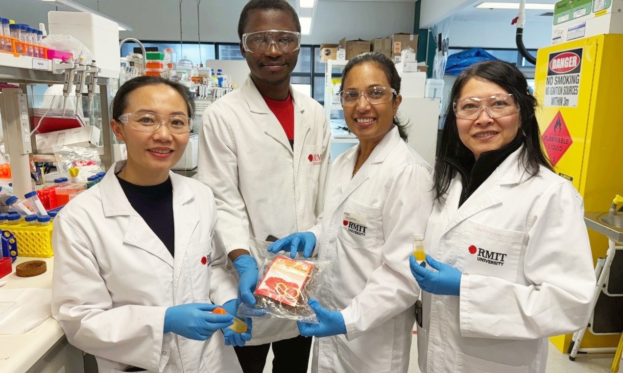 Four researchers in RMIT lab coats and blue gloves stand in a laboratory, holding small vials of orange lutein liquid and a packet of Gac fruit extract.
