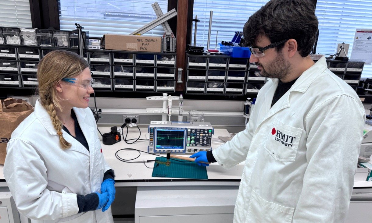 Two researchers in lab coats stand at a workbench next to electronic measurement equipment, with a nylon‑film sample positioned for testing using a hammer.