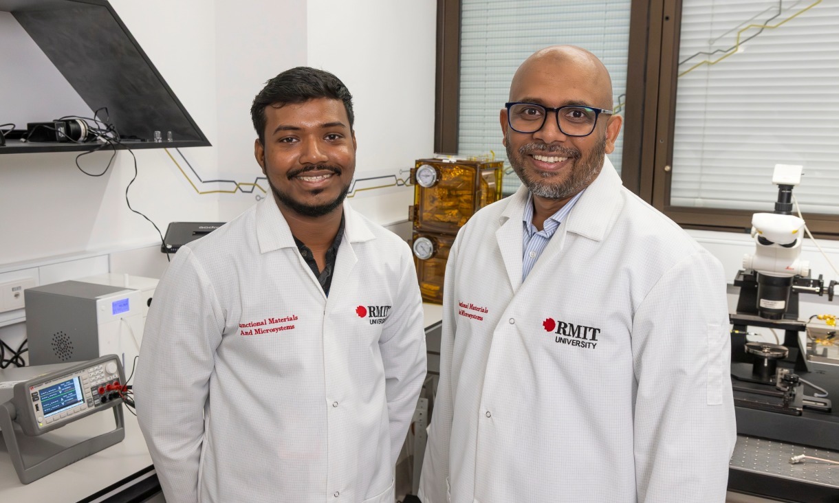 A small white robotic minibot shaped like a dolphin moves across the surface of a clear water tank, using a front‑mounted nozzle to collect a trail of blue kerosene oil floating on the water. The robot has a rounded body with a transparent dome on top and RMIT University branding on its side.