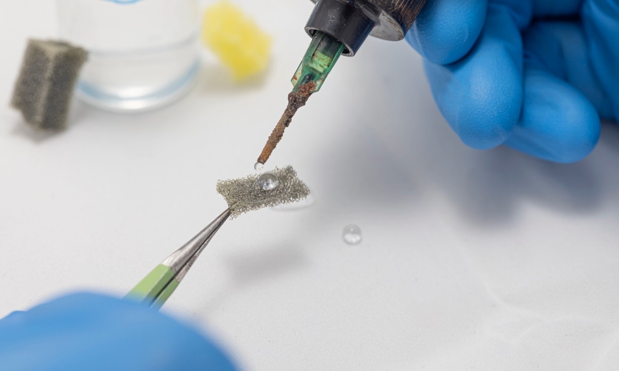 A gloved hand holds a small coated filter piece with tweezers while a droplet of water beads on the surface, demonstrating the material's water‑repellent behaviour.